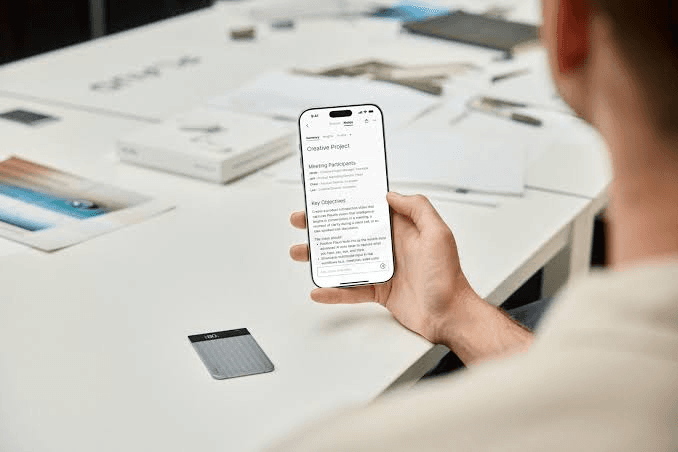 PLAUD Note on a desk beside a phone showing an AI meeting summary